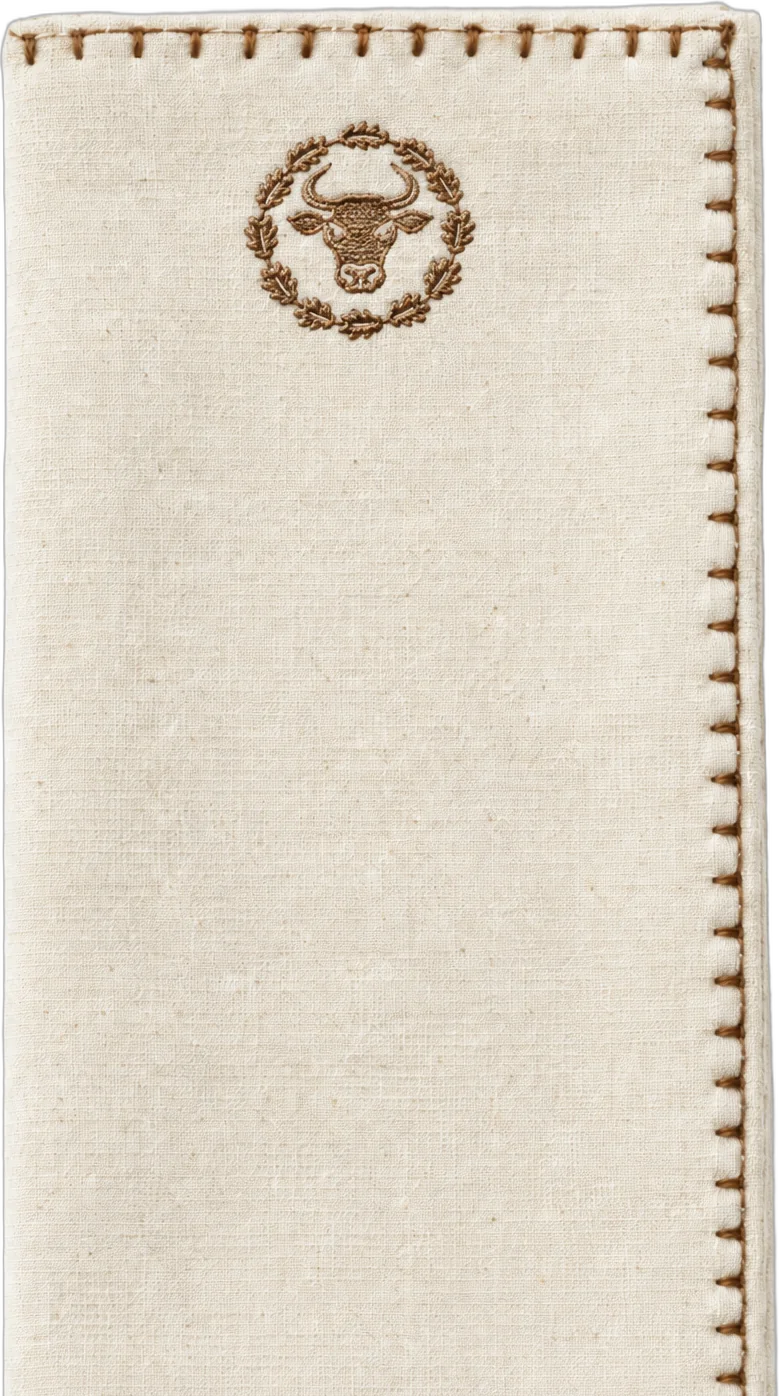 A folded cream cotton napkin with a hand-stitched brown border and the Steak Garden bull embroidered near the top